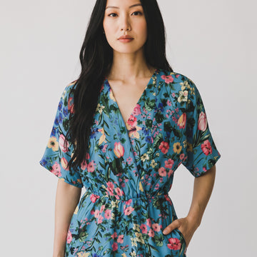 A woman wearing a blue short-sleeve wrap dress with a colorful floral print, standing against a plain light gray background.