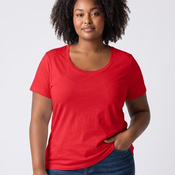 A woman with curly dark hair wearing a bright red short-sleeve scoop-neck t-shirt with a subtle tonal embroidered logo on the left chest, paired with blue jeans, against a light gray studio background.