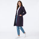 A woman modeling a long, dark purple tailored coat over a white top, blue jeans, and white sneakers, standing against a plain white studio background.