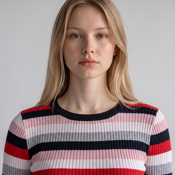 A close-up of a woman wearing a ribbed knit crewneck sweater with horizontal stripes in red, pink, gray, and navy blue against a light gray background.