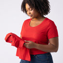 A woman wearing a bright red short-sleeve crewneck t-shirt with a small embroidered logo on the chest, holding a folded version of the t-shirt in her hands against a light gray background.