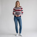 A woman stands against a light gray studio background wearing a fitted long-sleeve knit sweater with horizontal navy, red, white, and light gray stripes, blue jeans, and white sneakers.