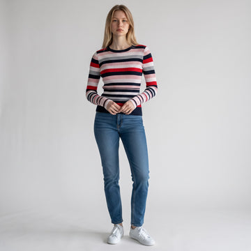 A woman stands against a light gray studio background wearing a fitted long-sleeve knit sweater with horizontal navy, red, white, and light gray stripes, blue jeans, and white sneakers.