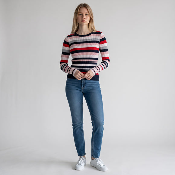 A woman stands against a light gray studio background wearing a fitted long-sleeve knit sweater with horizontal navy, red, white, and light gray stripes, blue jeans, and white sneakers.
