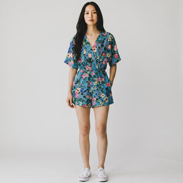 A woman modeling a short-sleeve floral romper with a V-neck and waist tie, paired with white sneakers, against a plain light gray studio background.