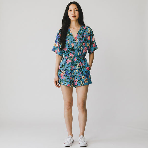 A woman modeling a short-sleeve floral romper with a V-neck and waist tie, paired with white sneakers, against a plain light gray studio background.