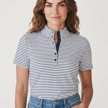 A woman wearing a short-sleeve polo shirt with navy blue and white horizontal stripes, featuring a split collar and a three-button placket, paired with blue jeans against a light gray studio background.