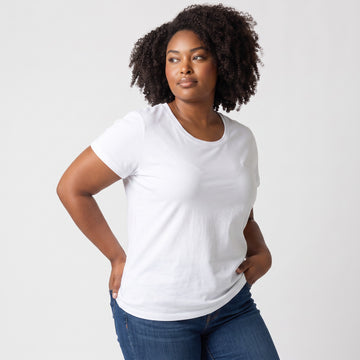 A woman with curly dark hair wearing a plain white short-sleeve crewneck t-shirt and blue jeans, posing against a light gray studio background.