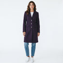 A woman modeling a dark purple knee-length coat with three front buttons, worn over a white top and blue jeans, against a plain white studio background.