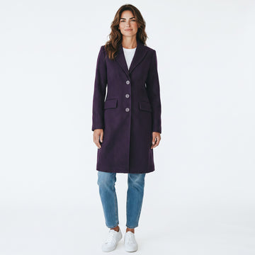 A woman modeling a dark purple knee-length coat with three front buttons, worn over a white top and blue jeans, against a plain white studio background.