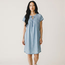 A woman modeling a light blue short-sleeve linen dress with a tie-neck detail, standing against a plain off-white background.