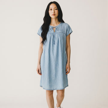 A woman modeling a light blue short-sleeve linen dress with a tie-neck detail, standing against a plain off-white background.