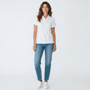 A woman modeling a white short-sleeve polo shirt with light blue jeans and white sneakers, standing against a plain white studio background.