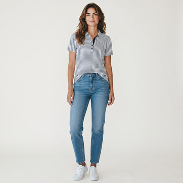 A woman modeling a short-sleeve polo shirt with thin navy and white horizontal stripes, paired with light-wash blue jeans and white sneakers, against a plain white studio background.